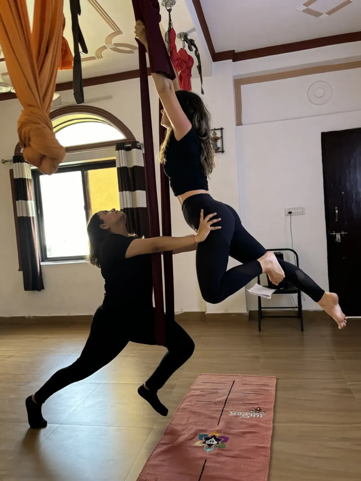 Aerial yoga teacher supporting a student's legs as she pulls herself up into the aerial silk