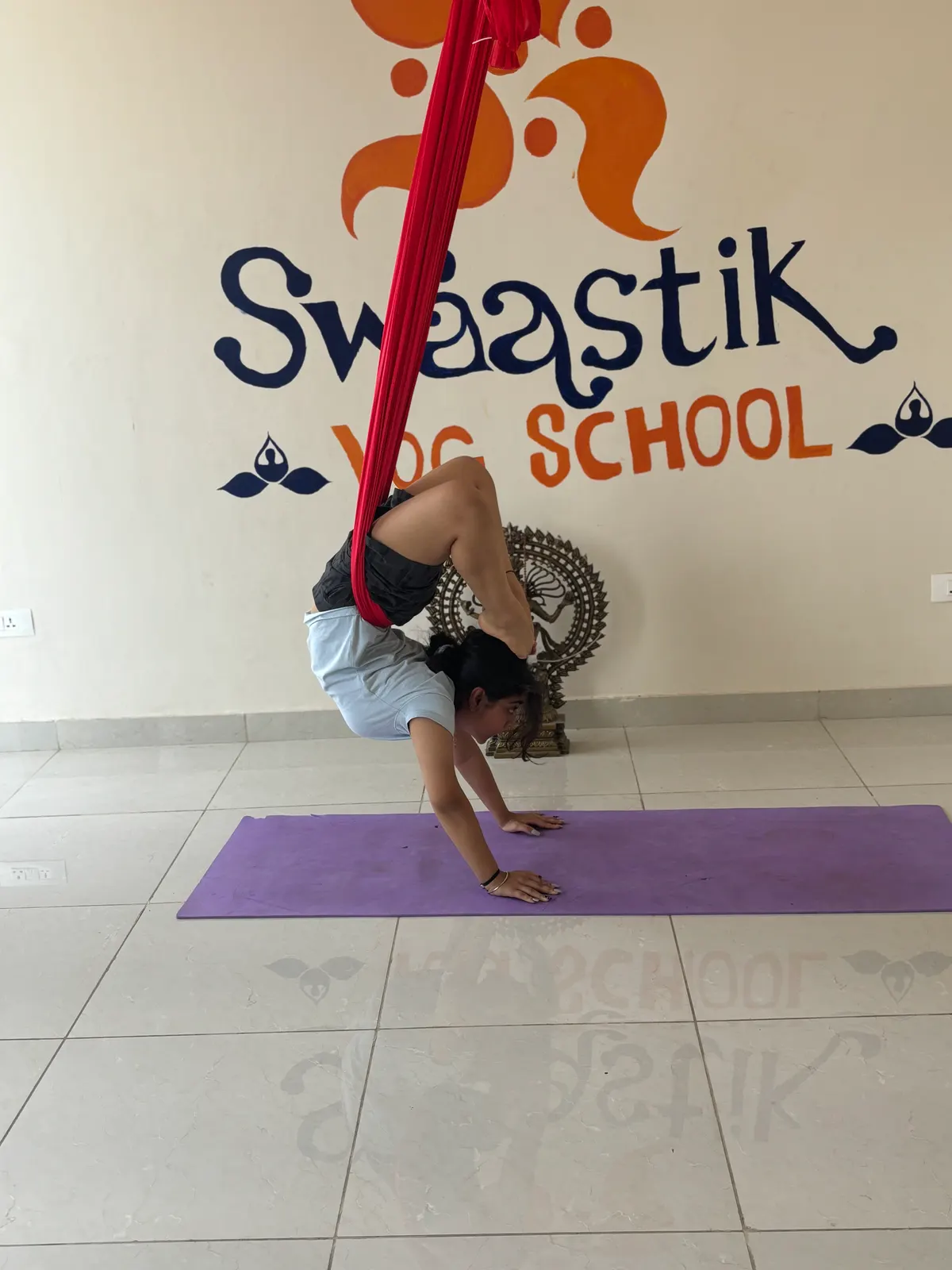 Student kicking up into a handstand with a red aerial hammock under her hips, beneath the painted Swaastik Yog School wall logo