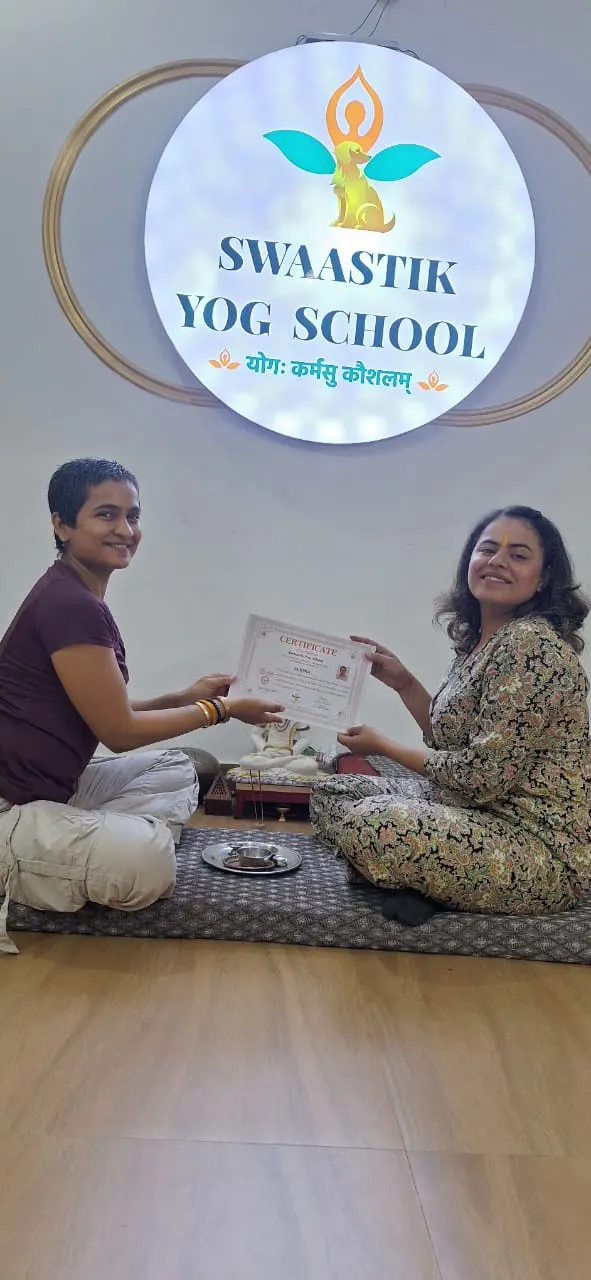 Teacher handing an aerial yoga teacher training certificate to a seated graduate beneath the Swaastik Yog School logo