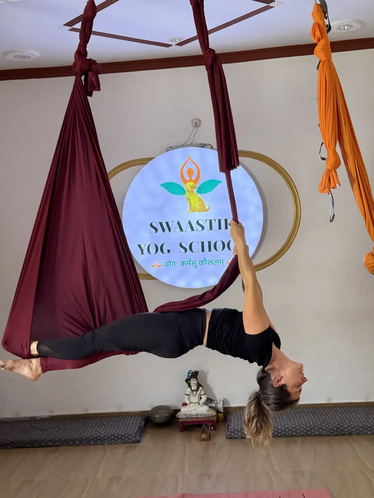 Student hanging upside down in an inverted aerial backbend, feet hooked in a maroon hammock, during aerial yoga teacher training in Rishikesh