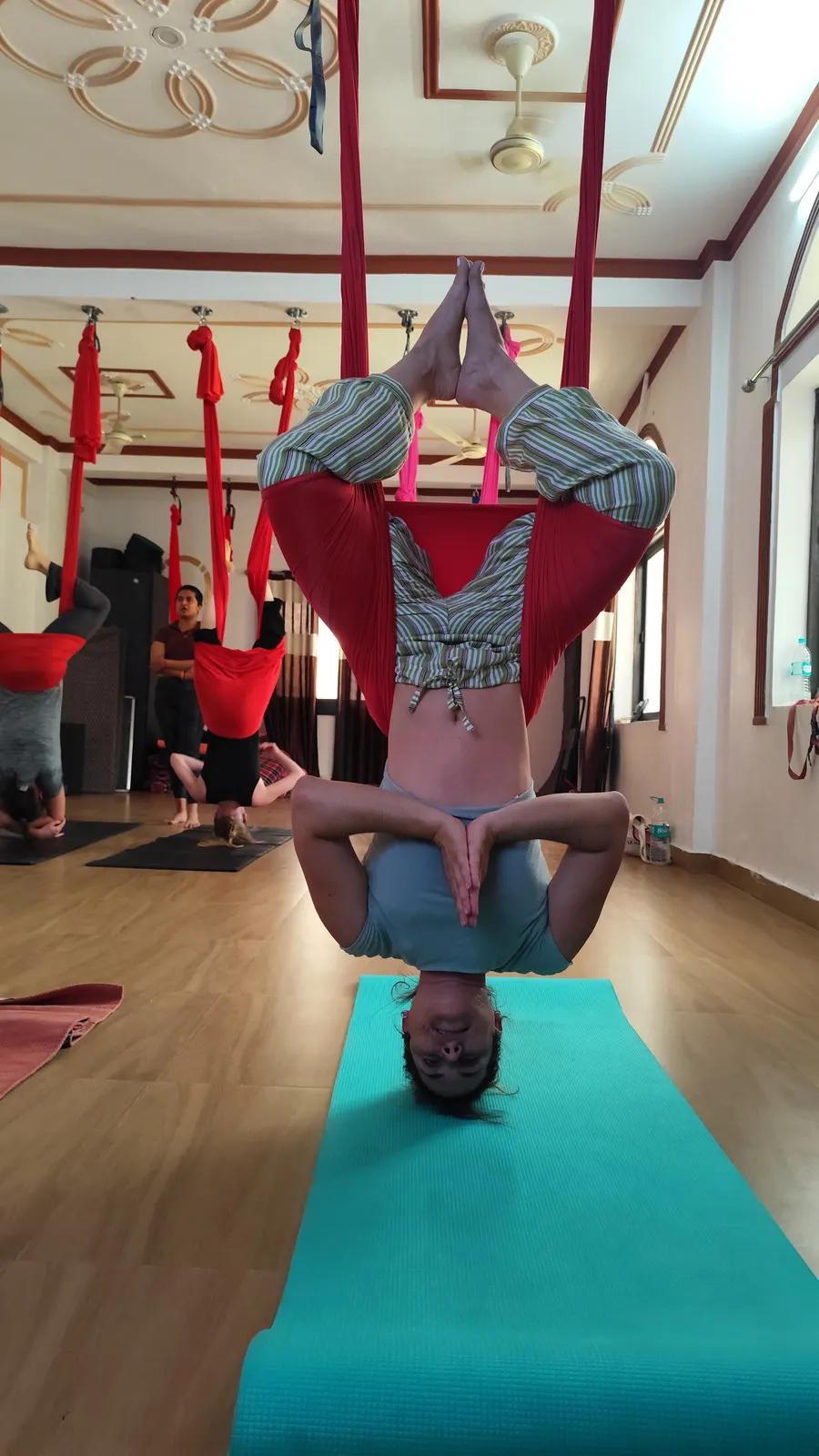 Student hanging upside down in inverted lotus, legs crossed in a red hammock and hands in prayer, during an aerial yoga class