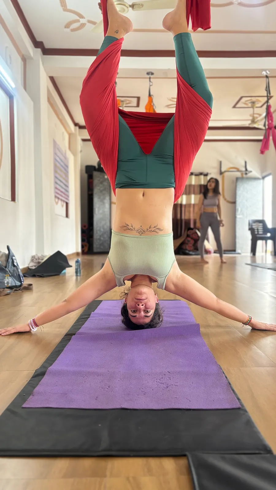 Student hanging fully inverted in a red aerial hammock with arms spread wide above a purple yoga mat