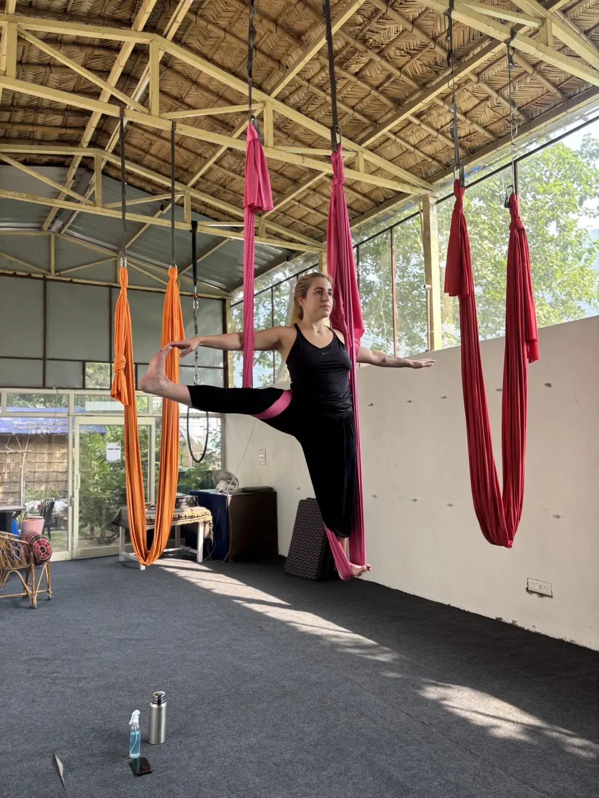 Student balancing in an aerial standing split with a pink hammock under her hip in the open-air bamboo-roof aerial shala in Rishikesh