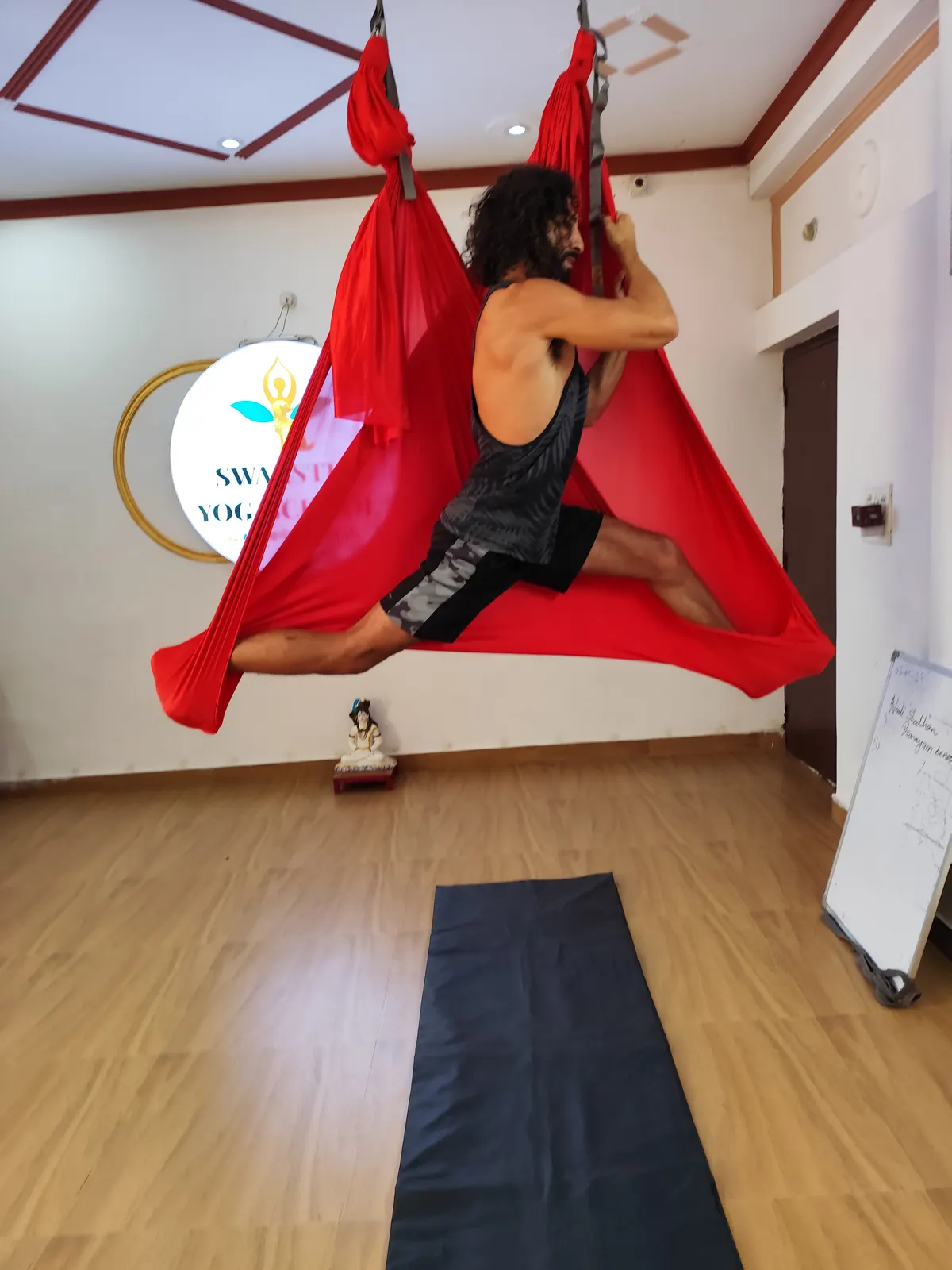 Student sitting in a wide aerial straddle inside a red hammock with legs extended, in the aerial yoga studio