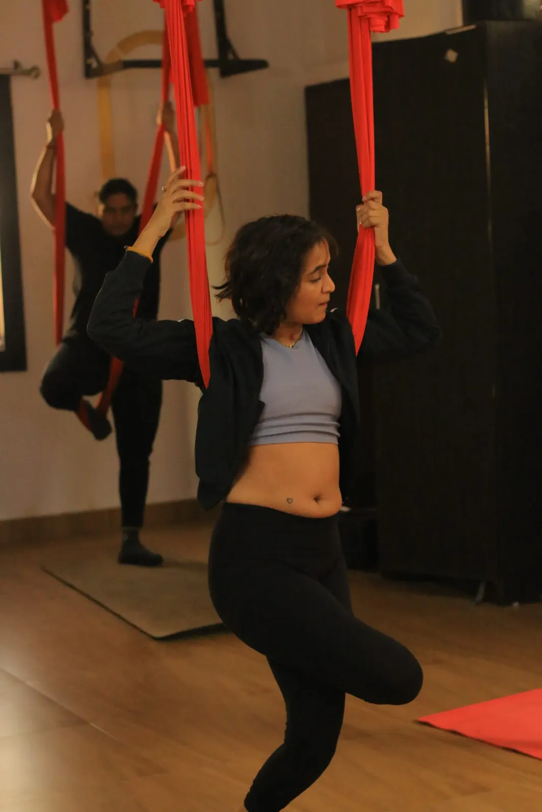 Student balancing in tree pose while holding two red aerial silks, with another student practising behind her